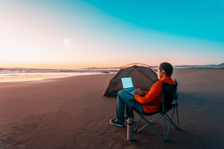Digital nomad working on a laptop at the beach during sunrise, balancing travel and work while camping.